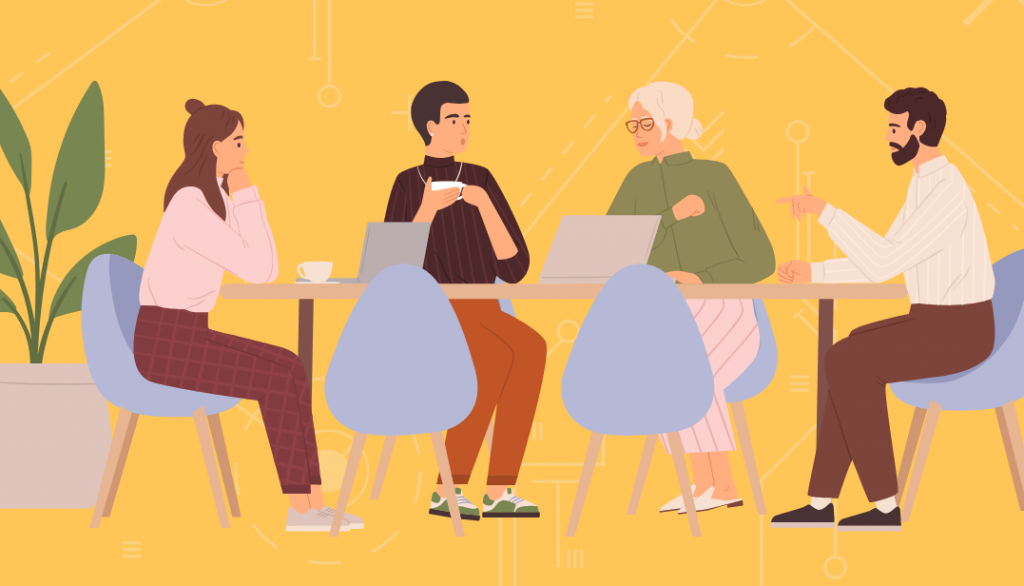 A group of four people sitting around a table, having a conversation. They have laptops and coffee mugs.