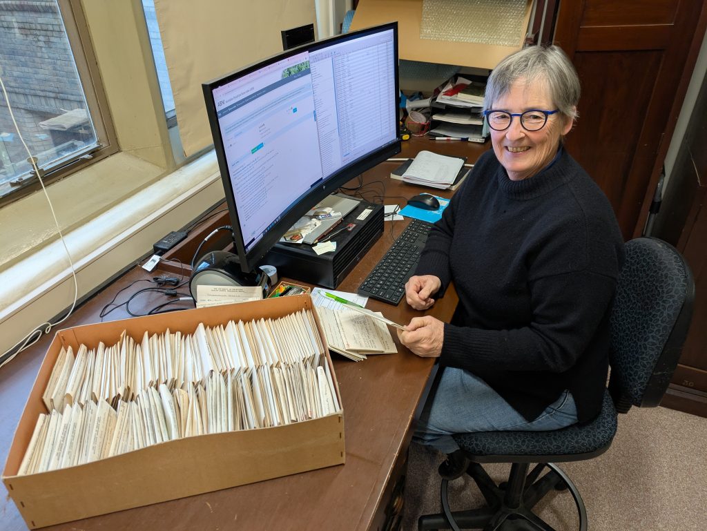 A person smiling, sitting at a desk with computer and large tray of papers ready to be digitised